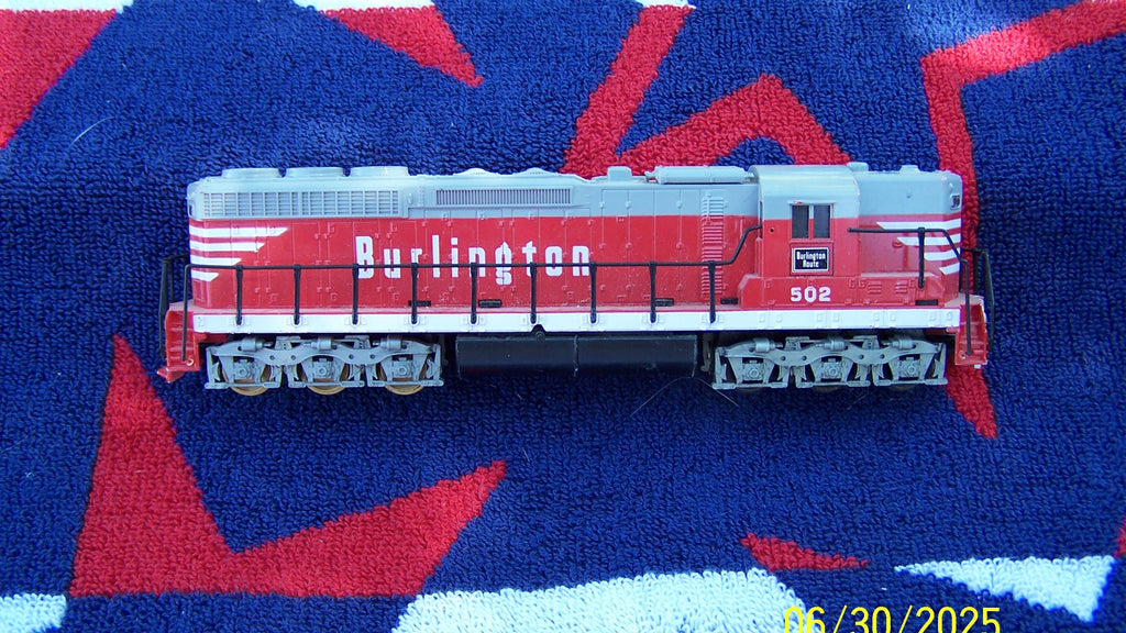 A red and gray model Burlington locomotive train, numbered 502, resting on a textured blue, red, and white blanket. The image has a yellow digital timestamp in the bottom right corner.